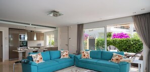 A modern living room with a teal sofa, a kitchen area, and a large sliding glass door leading to a balcony with greenery. - NEW!!! Beachfront IV, Pool, Garden, and Luxury Home, Elviria, Marbella. (Marbella)