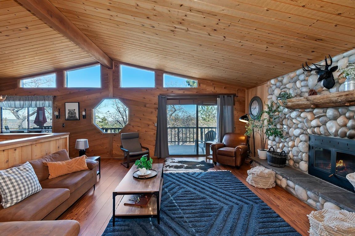 A cozy cabin interior with a stone fireplace, wooden ceiling, and a large window overlooking a deck.