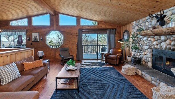 A cozy cabin interior with a wooden ceiling, a stone fireplace, a large window, and a comfortable seating area. - Close to Bear Mountain Ski Resort, Golf Course and Zoo! PET FRIENDLY 🐾! Rock Fireplace! Mountain Views! (Big Bear City)