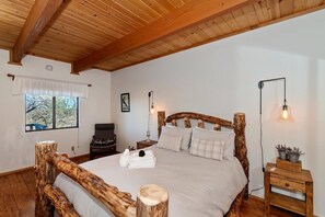 A bedroom with a wooden headboard, a bed with white linens, a nightstand with a lamp, and a window with a view of trees. - Close to Bear Mountain Ski Resort, Golf Course and Zoo! PET FRIENDLY 🐾! Rock Fireplace! Mountain Views! (Big Bear City)