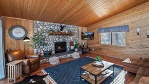 A cozy log cabin interior with a stone fireplace, leather sofas, a coffee table, and a television mounted on the wall. - Close to Bear Mountain Ski Resort, Golf Course and Zoo! PET FRIENDLY 🐾! Rock Fireplace! Mountain Views! (Big Bear City)