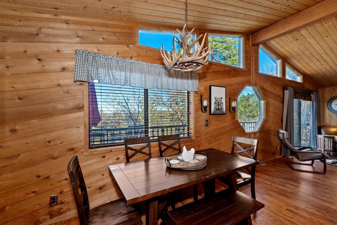 A wooden interior with a dining table, chairs, a chandelier, and large windows.