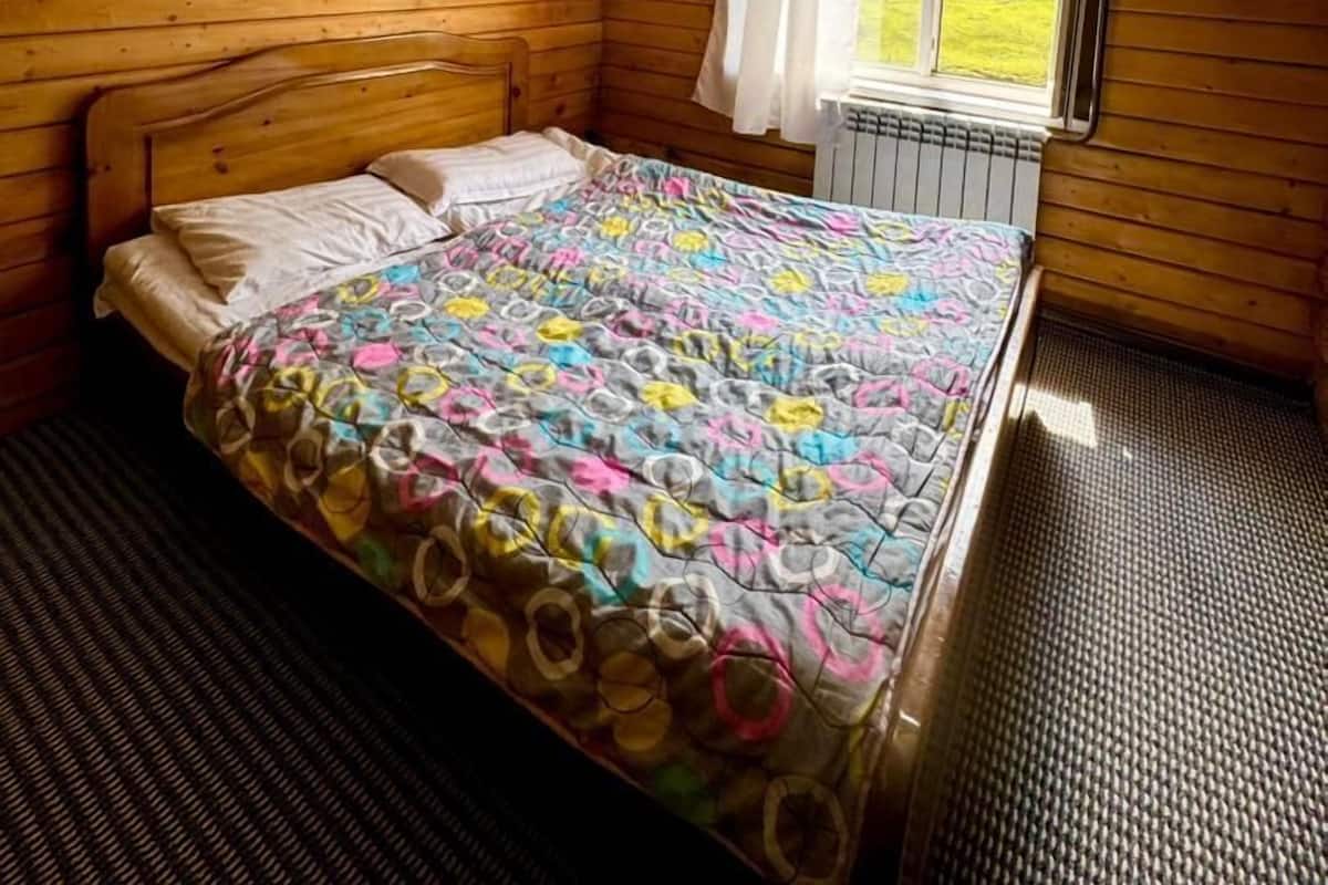 A wooden cabin bedroom with a bed, a window with curtains, and a view of a forest.