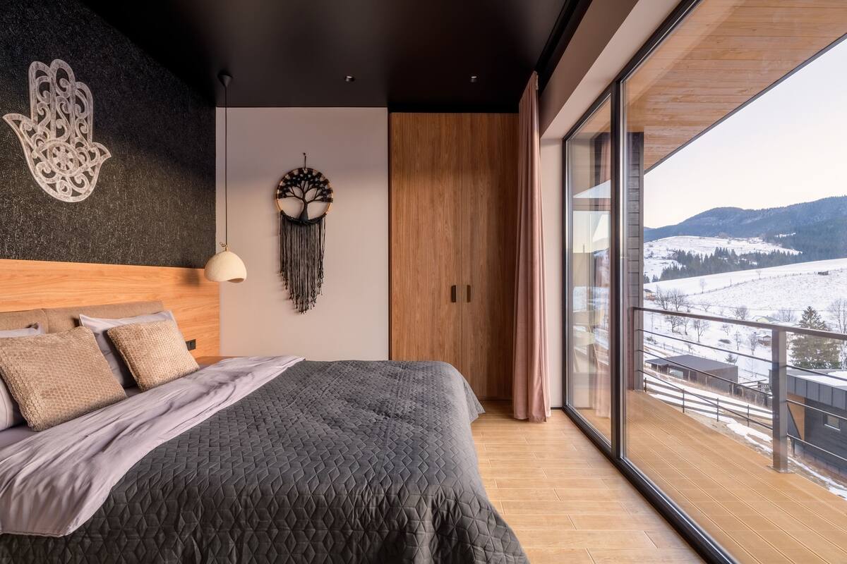 Une chambre moderne avec un grand lit, un attrape-rêves accroché au mur et une vue sur un paysage enneigé par de grandes fenêtres.