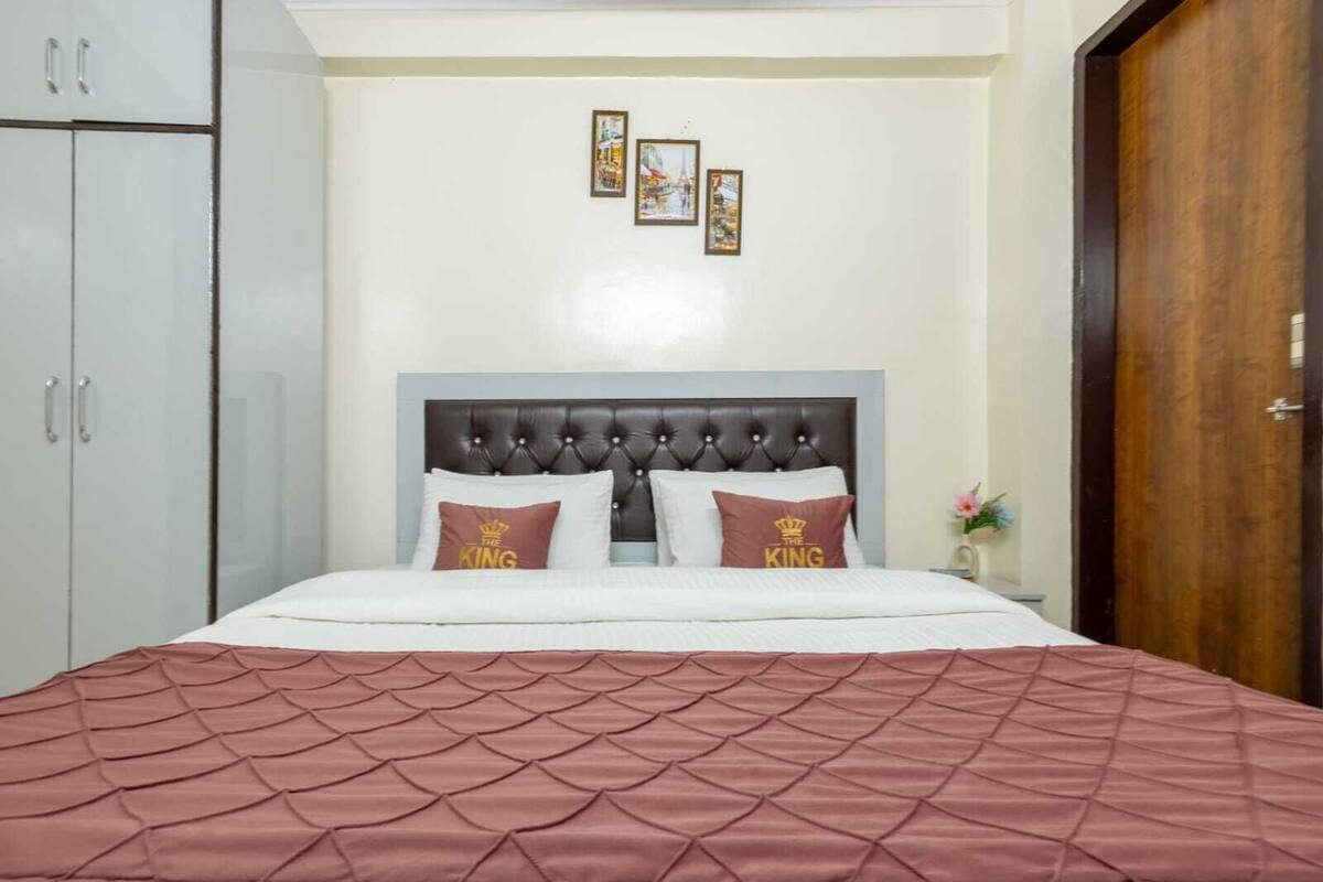 A bed with a tufted headboard, two pillows with 'THE KING' text, a pink quilted bedspread, and a wall with three framed pictures.
