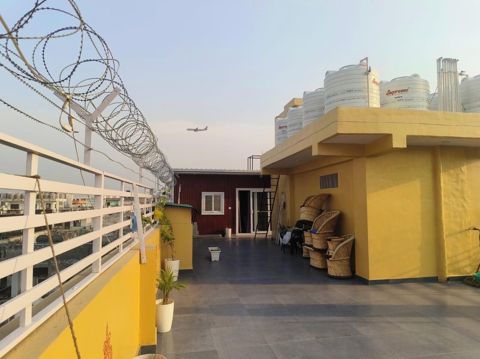 A rooftop with a barbed wire fence, a red building, and stacked white containers.