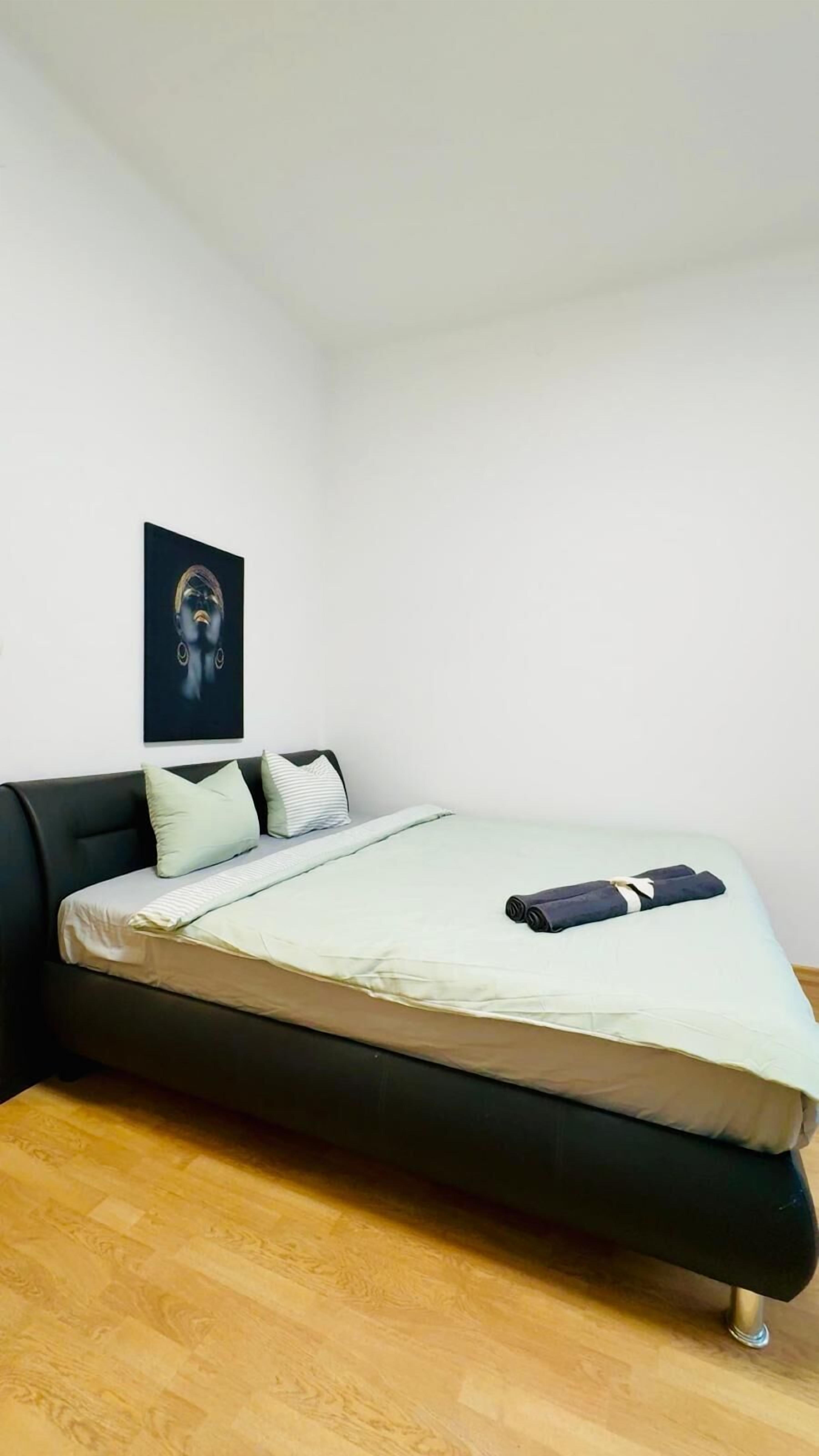 Een enkel bed met een zwart frame, een witte matras en een lichtkleurige deken. Er ligt een kussen met een gestreept patroon en een kussen in effen kleur op. Een donkere, opgerolde deken ligt op het bed. De kamer heeft een houten vloer en een witte muur.