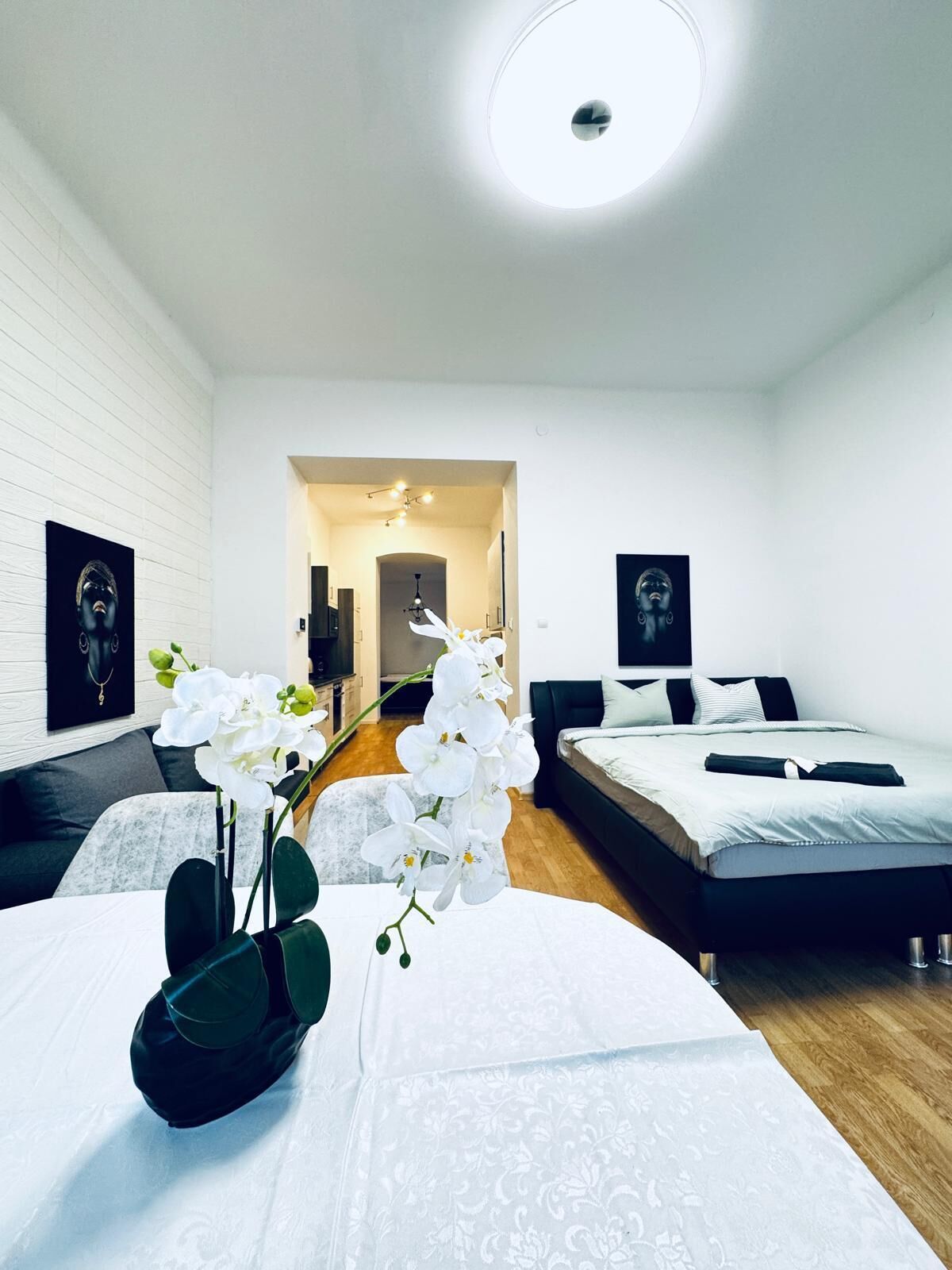 A modern hotel room with a white brick wall, a round ceiling light, two beds with white bedding, a small table with a vase of white orchids, and a hallway leading to a bathroom.