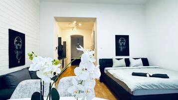 A modern hotel room with a white brick wall, a round ceiling light, two beds with white bedding, a small table with a vase of white orchids, and a hallway leading to a bathroom.