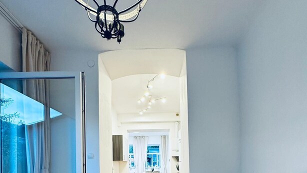 A bedroom with a bed, a window with curtains, a chandelier, and a view of a hallway leading to a bathroom. - Lovely 2BR for 5 Peaceful Central Stay (Klosterneuburg)
