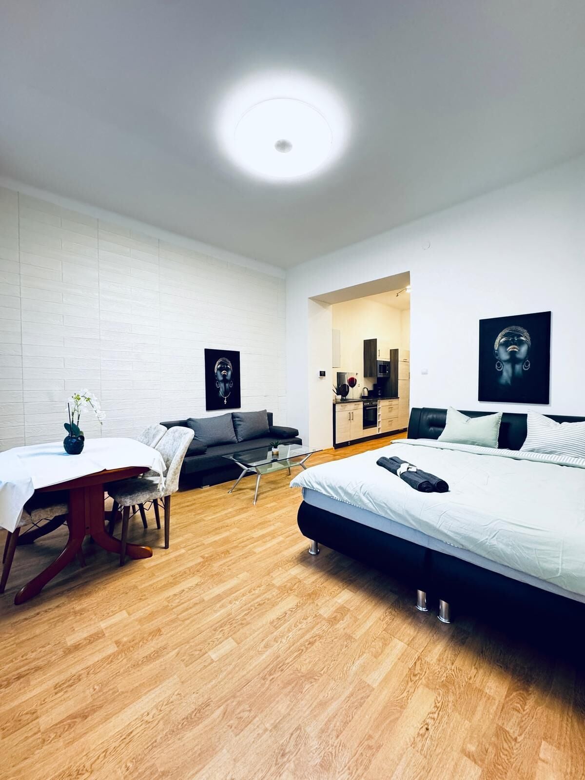 A modern hotel room with a wooden floor, a bed, a dining table with chairs, a sofa, and a kitchenette.