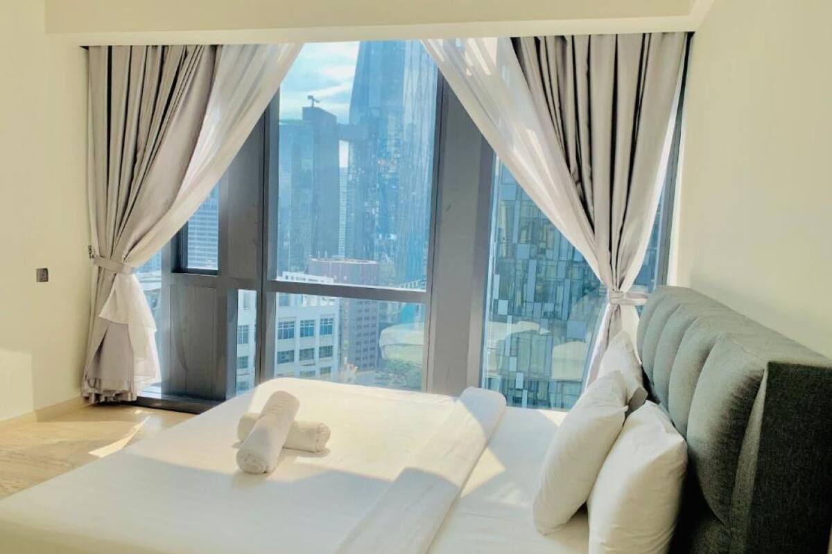 A modern hotel room with a large bed, white linens, and a view of a cityscape through floor-to-ceiling windows.