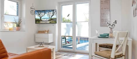 A bright, open living space with a sofa, a dining table with chairs, a TV mounted on the wall, and a large window offering a view of the outdoors.