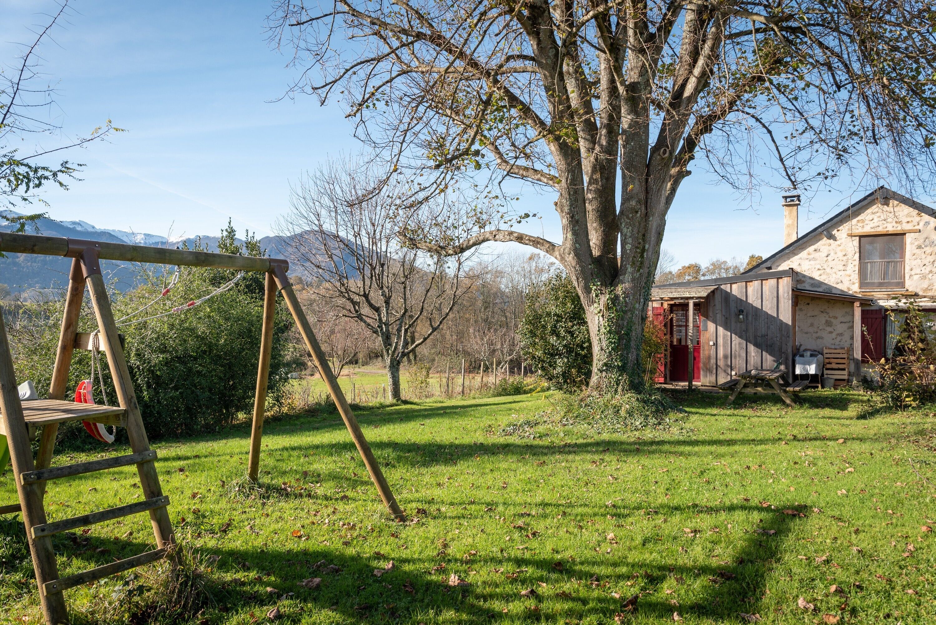 A wooden swing set, a large tree, a stone building with a red door, and a mountain in the background.