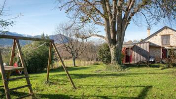 A wooden swing set, a large tree, a stone building with a red door, and a mountain in the background.