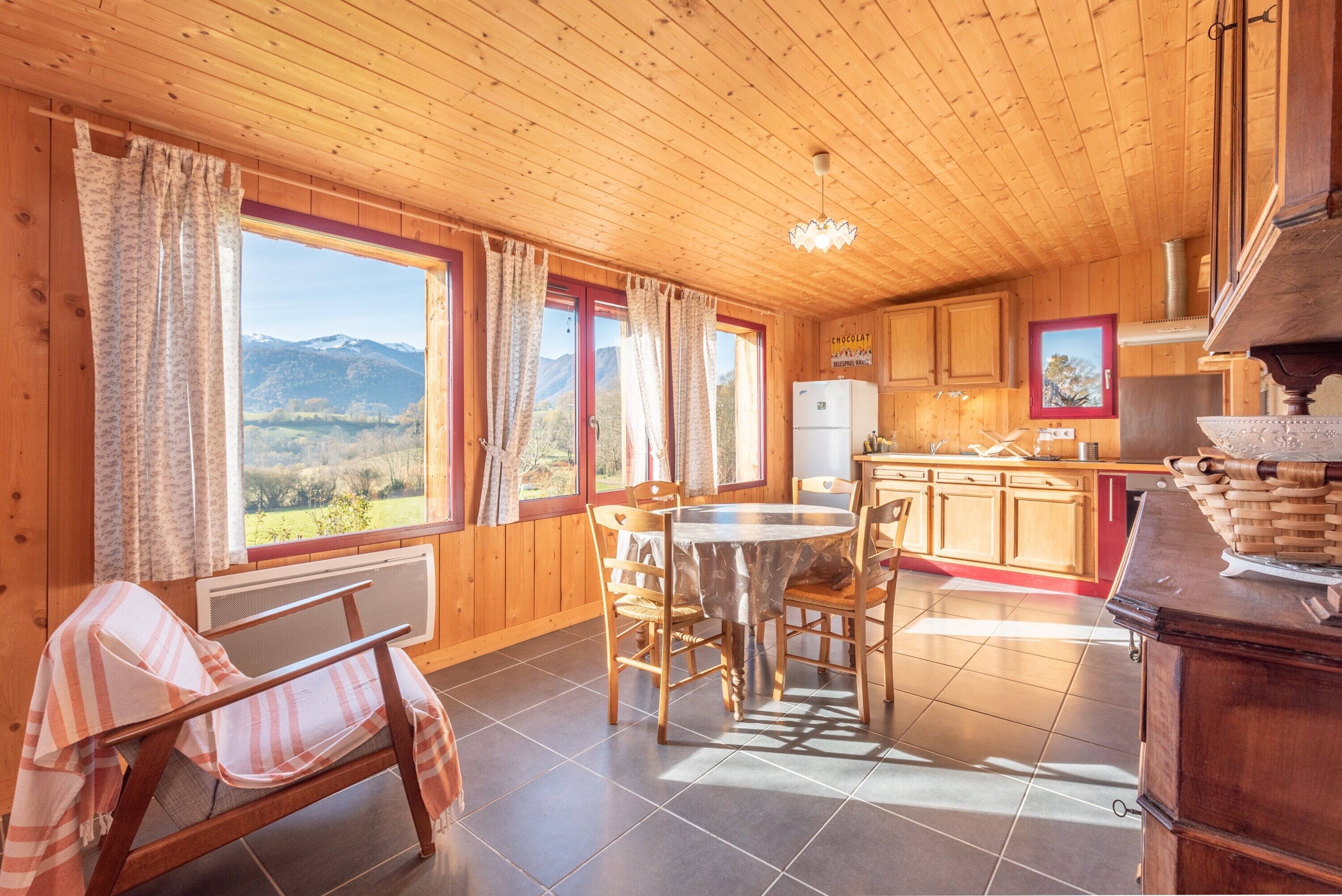 L’intérieur d’un chalet en bois comprend une table à manger, des chaises, un coin cuisine et de grandes fenêtres offrant une vue sur la montagne.