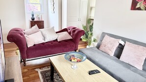 A living room with a maroon sofa, a grey sofa, a coffee table, and a TV. There is a cabinet, a plant, and a window with blinds.