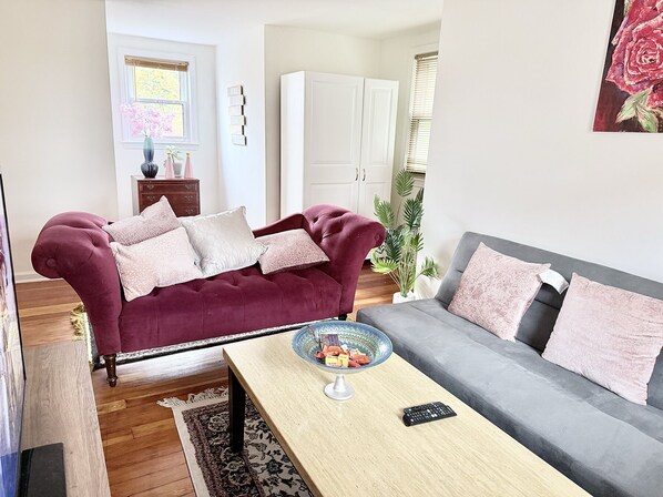 Ett vardagsrum med en vinröd soffa, en grå soffa, ett soffbord och en tv. Det finns ett skåp, en växt och ett fönster med persienner.