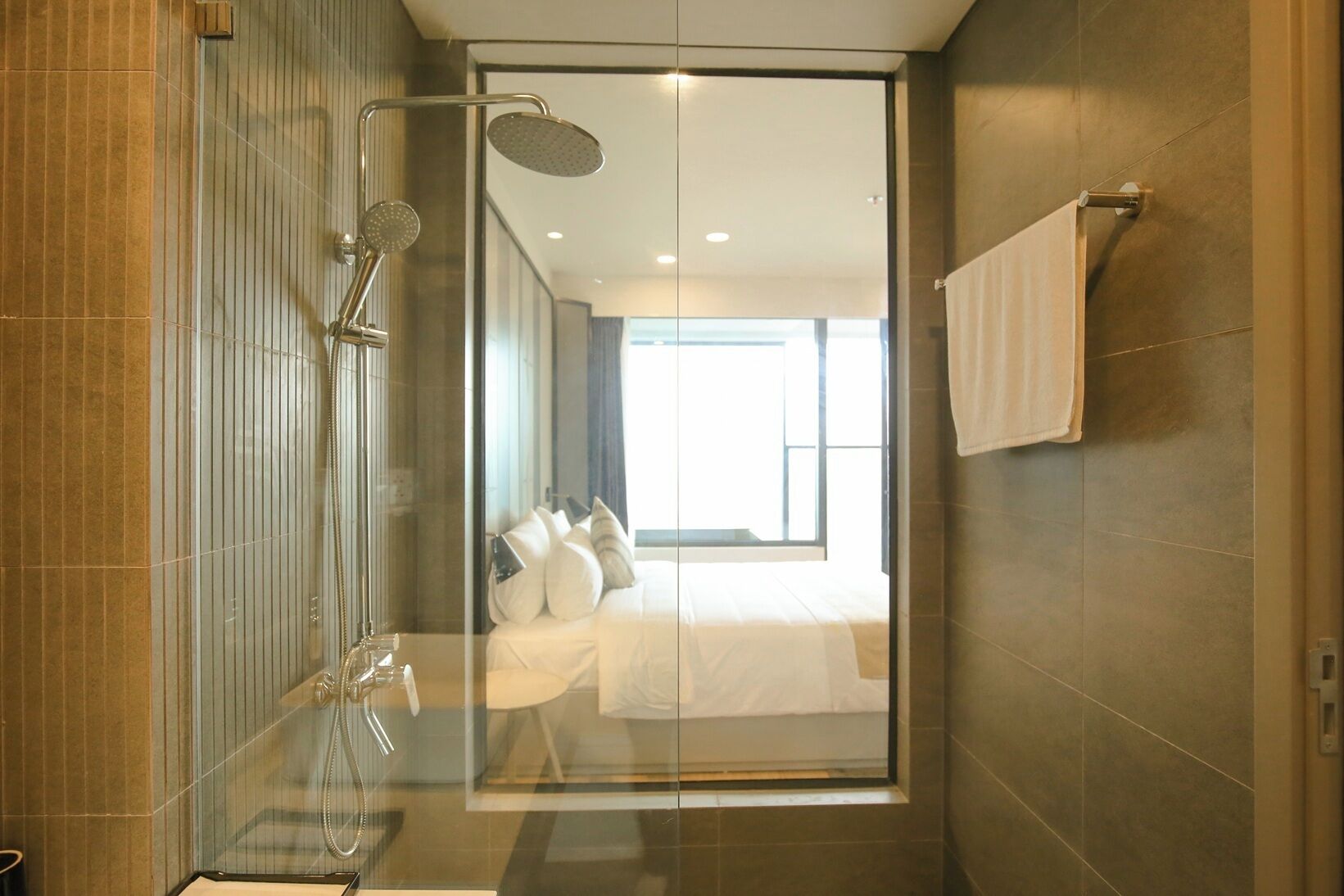 A modern bathroom with a glass shower enclosure, a visible bed with white bedding, and a towel rack on the wall.