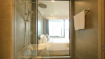 A modern bathroom with a glass shower enclosure, a visible bed with white bedding, and a towel rack on the wall.