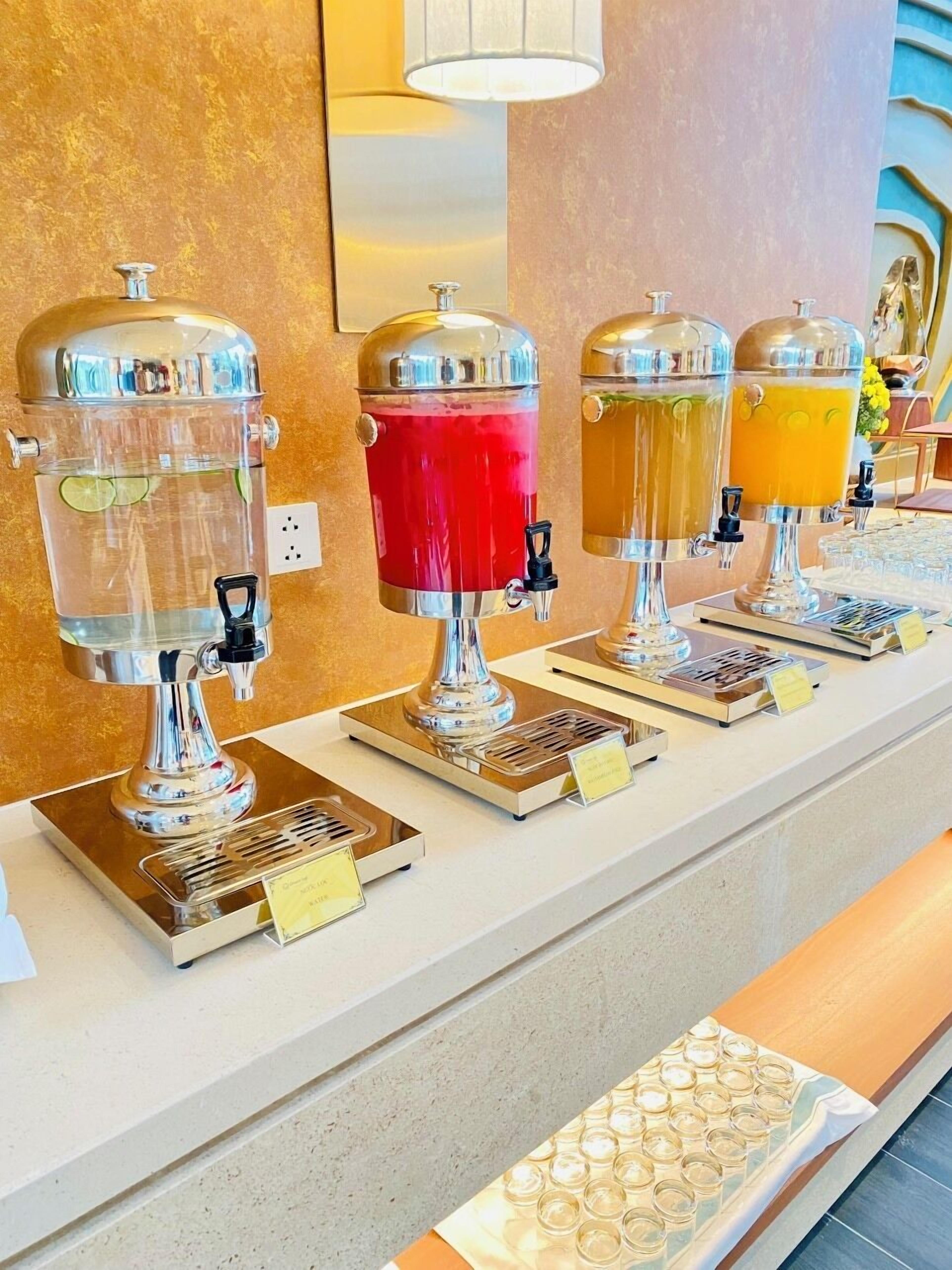 Una estación de bebidas con cuatro dispensadores, cada uno con tapa de color diferente, y una bandeja de vasos debajo.