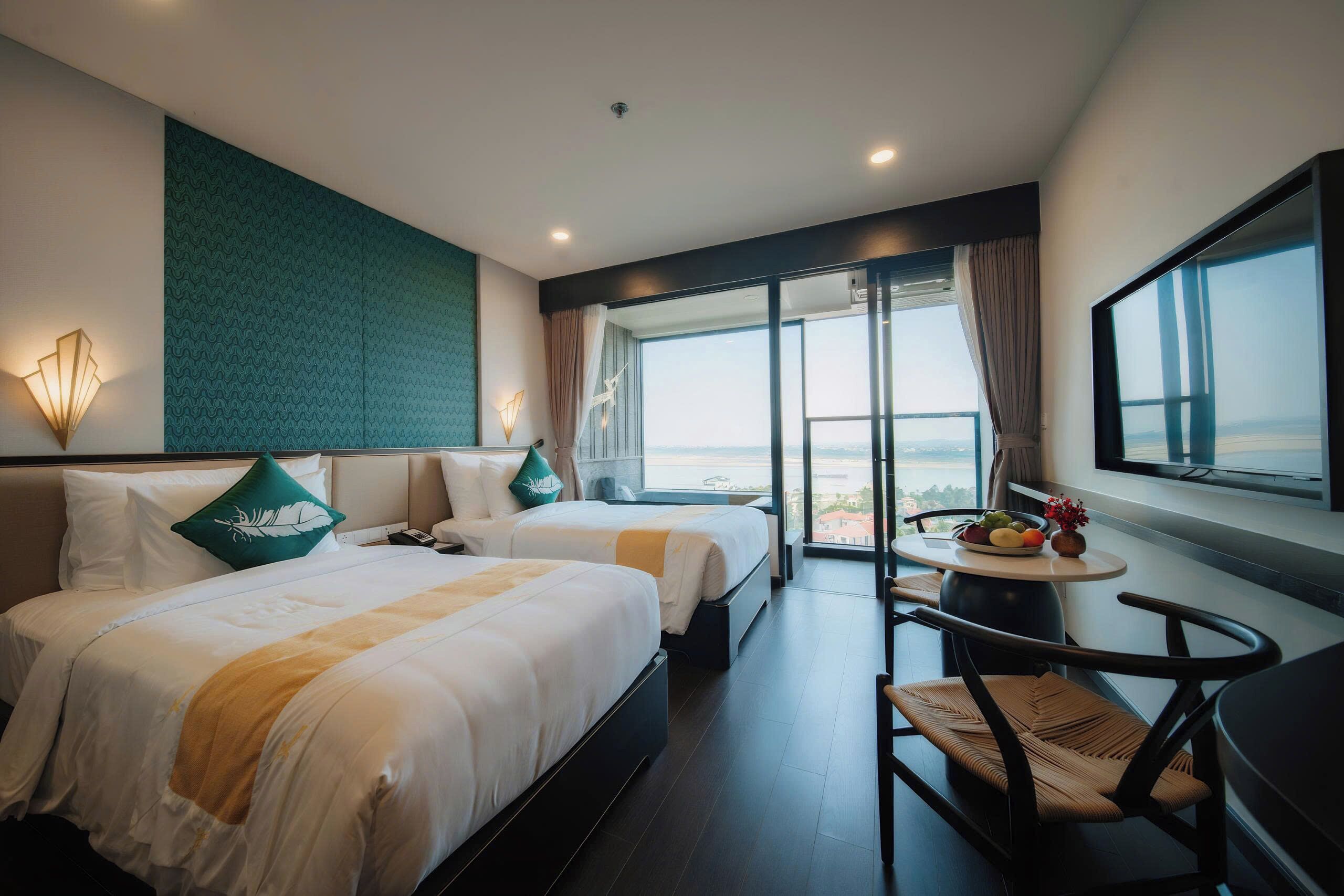 A hotel room with two beds, a dining table with fruit, a chair, and a large window with a view.