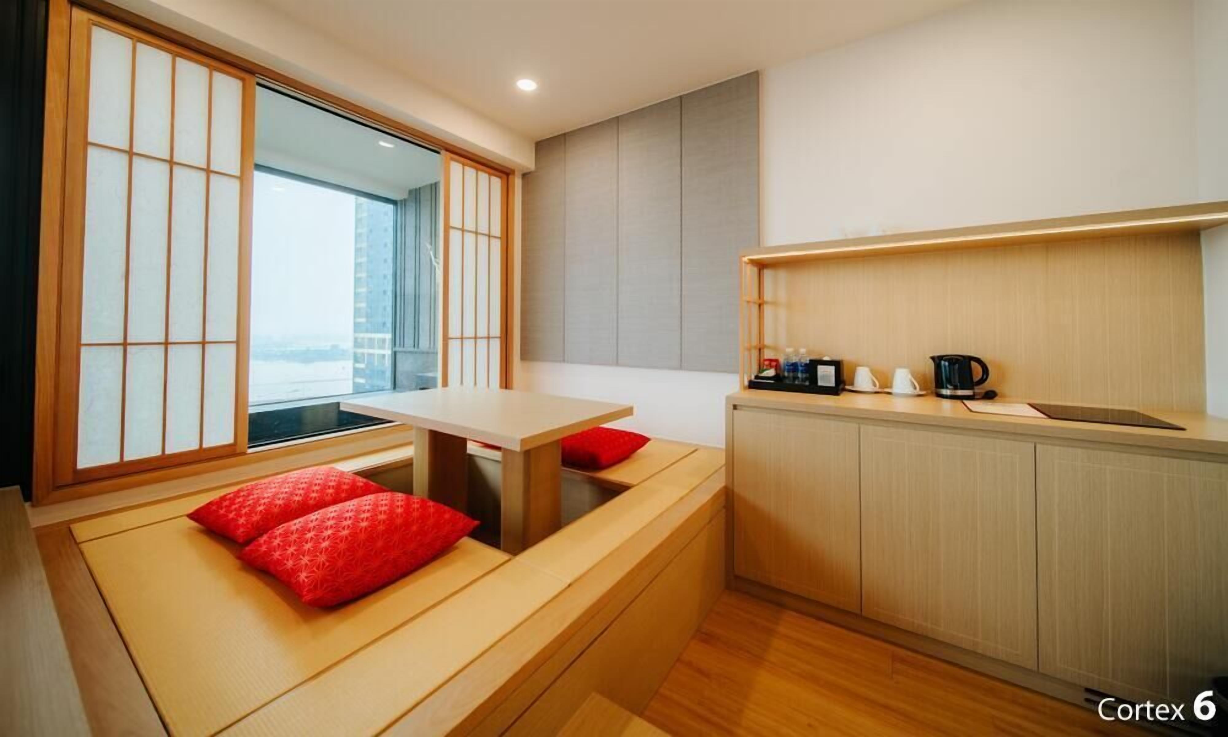 A compact hotel room with a kitchenette, a dining area with a low table and benches, and a large window offering a view of the city.