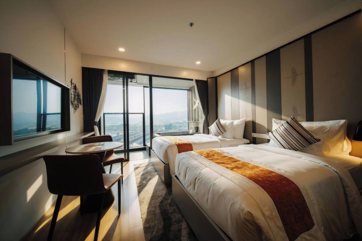 Ein modernes Hotelzimmer mit zwei Betten, einem Schreibtisch und einem großen Fenster mit Blick auf die Stadt.