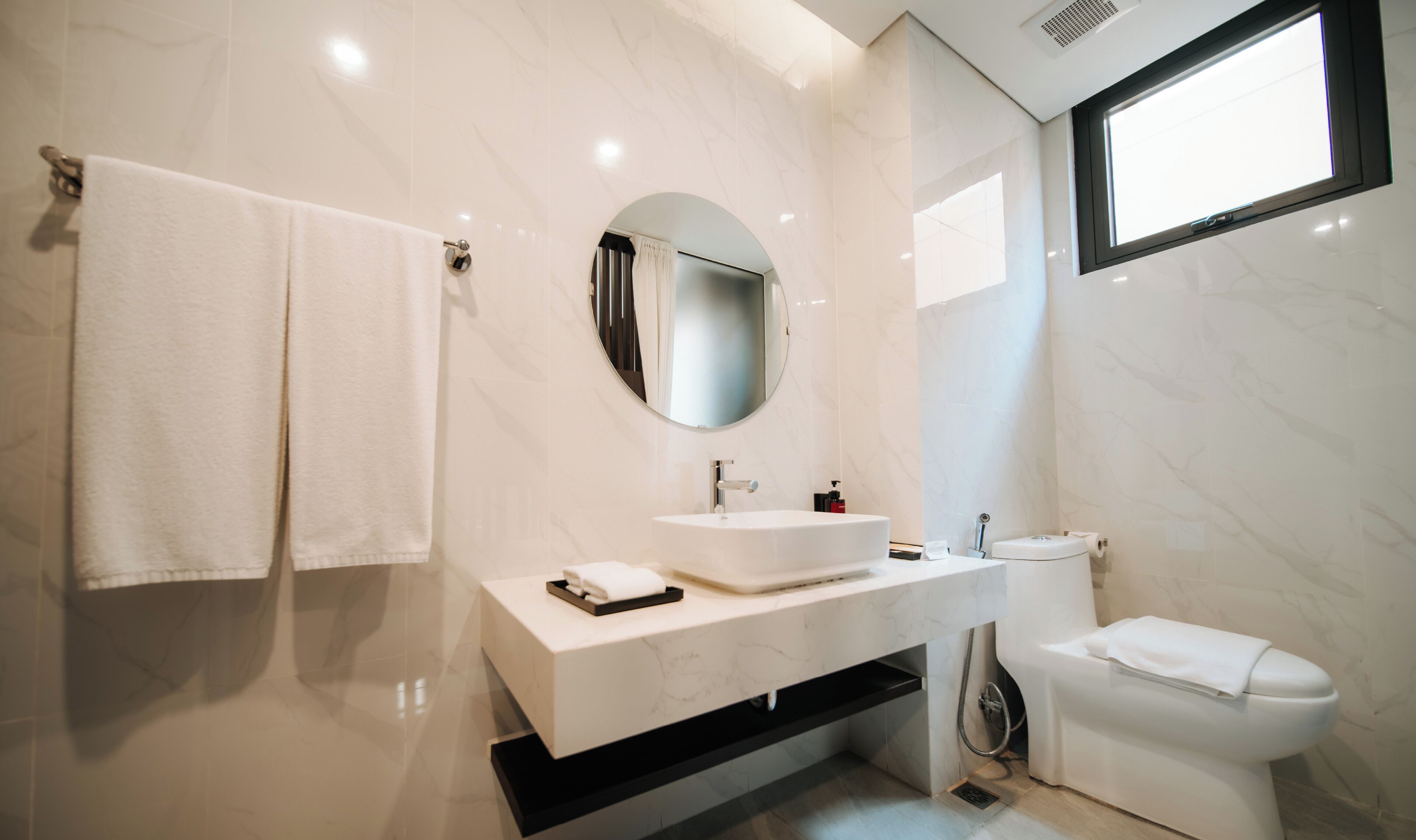 A modern bathroom with a white vanity, a round mirror, a toilet, and a window.