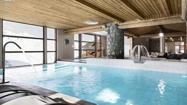 Piscina interna con un pilastro in pietra, travi in legno a soffitto e vista sulle montagne attraverso ampie finestre.