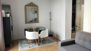 Een knus woongedeelte met een ronde glazen tafel, twee witte stoelen, een grijze bank, een grote spiegel en een vloerlamp.