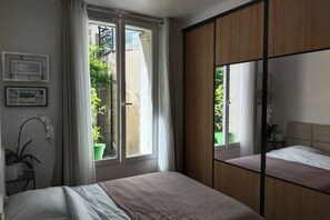 A bedroom with a bed, a sliding wooden wardrobe, a window with curtains, and a potted plant. - Charming 2-room apartment, quiet location (classified as furnished) (Boulogne-Billancourt)
