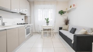 A modern kitchen with white cabinets, a dining table with chairs, a black sofa, and a window with sheer curtains. - "La Casa di Mia" Apartment – Cagliari (Cagliari)