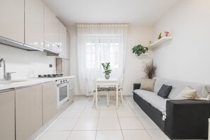 Una cucina moderna con mobili bianchi, un tavolo da pranzo con sedie, un divano nero e una finestra con tende leggere.
