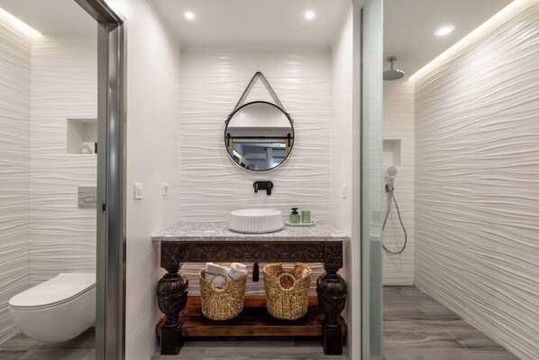 Un baño moderno con espejo redondo, paredes blancas con textura, tocador de madera y cabina de ducha de vidrio.