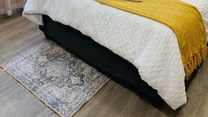 Un lit bien fait, avec du linge de lit blanc et un plaid jaune, une lampe de chevet et un tapis à motifs sur un plancher en bois.