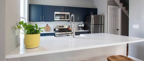 Una cocina moderna con armarios azul oscuro, electrodomésticos de acero inoxidable, una isla blanca con taburetes e iluminación colgante.