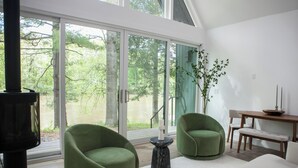 A modern living room with a large sliding glass door leading to a forested area, two green armchairs, a white sofa, a small wooden table, and a black heater. - Cozy Cabin On The Esopus Creek in Saugerties, New York (Saugerties)