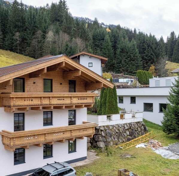 Un chalet à deux étages doté d’un balcon en bois, d’un mur de soutènement en pierre et offrant une vue sur une colline boisée.