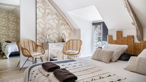 Une chambre avec un plafond en pente, un lit avec des oreillers et une couverture, deux fauteuils en osier, une commode et une fenêtre.