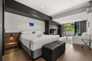 Una habitación de hotel moderna con una cama grande, una zona de estar con sofá y sillón, una mesita y un balcón con vistas a zonas verdes.