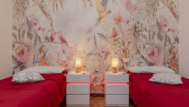 Ein Zimmer mit zwei Betten, jeweils mit roter Bettdecke und einem gemusterten Kissen. An beiden Betten stehen Nachttische mit Lampen, und an der Wand ist ein Wandbild mit Vögeln und Blumen zu sehen.
