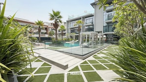 A modern outdoor pool area with lounge chairs, palm trees, and a glass-enclosed cabana. - Manzil - 1BR Apartment in JVC with Pool  (Dubai)