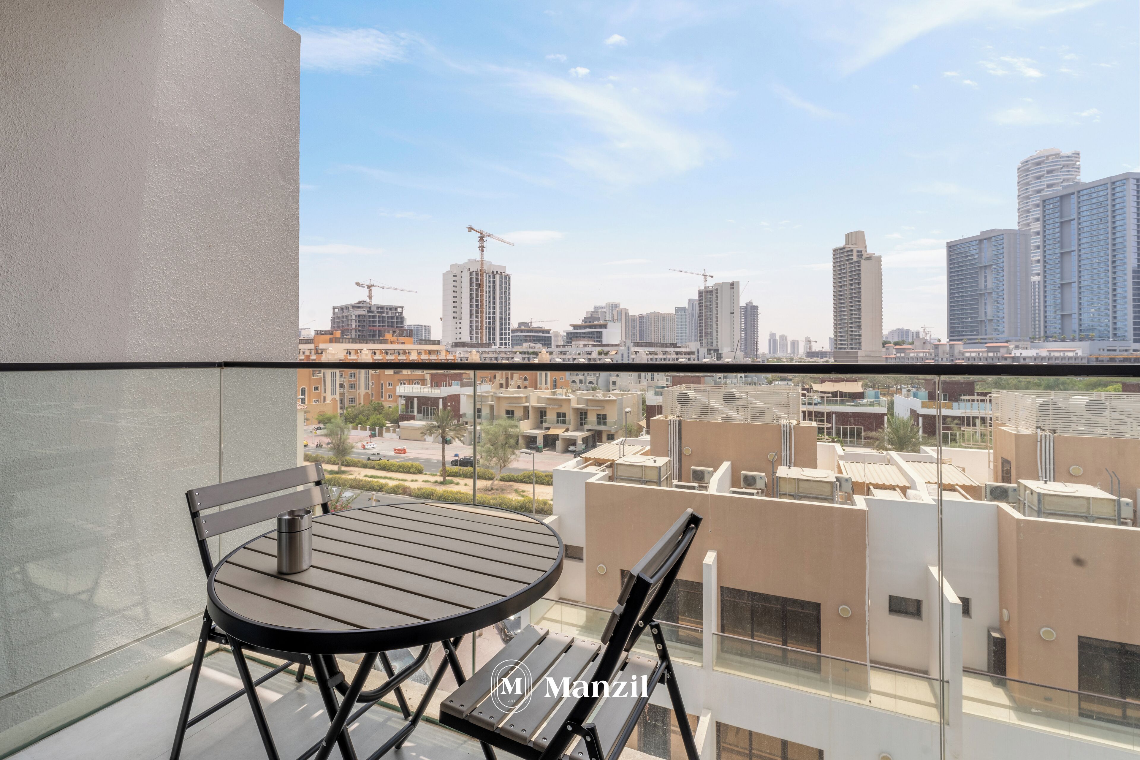 A balcony with a table and chairs, overlooking a cityscape with buildings and construction cranes.