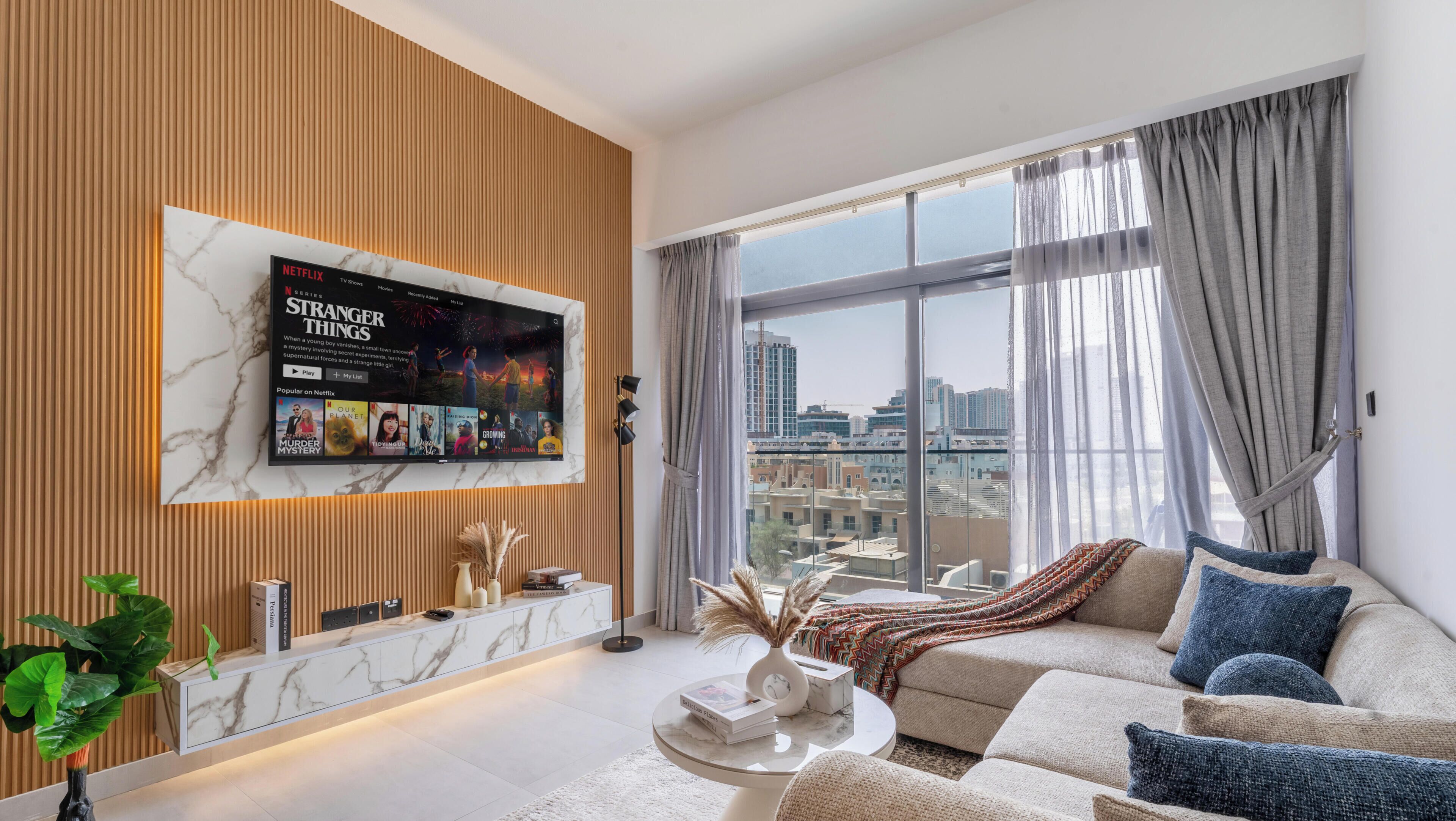 A modern living room with a large TV mounted on a wooden wall, a sofa, a coffee table, and a view of the city through large windows.