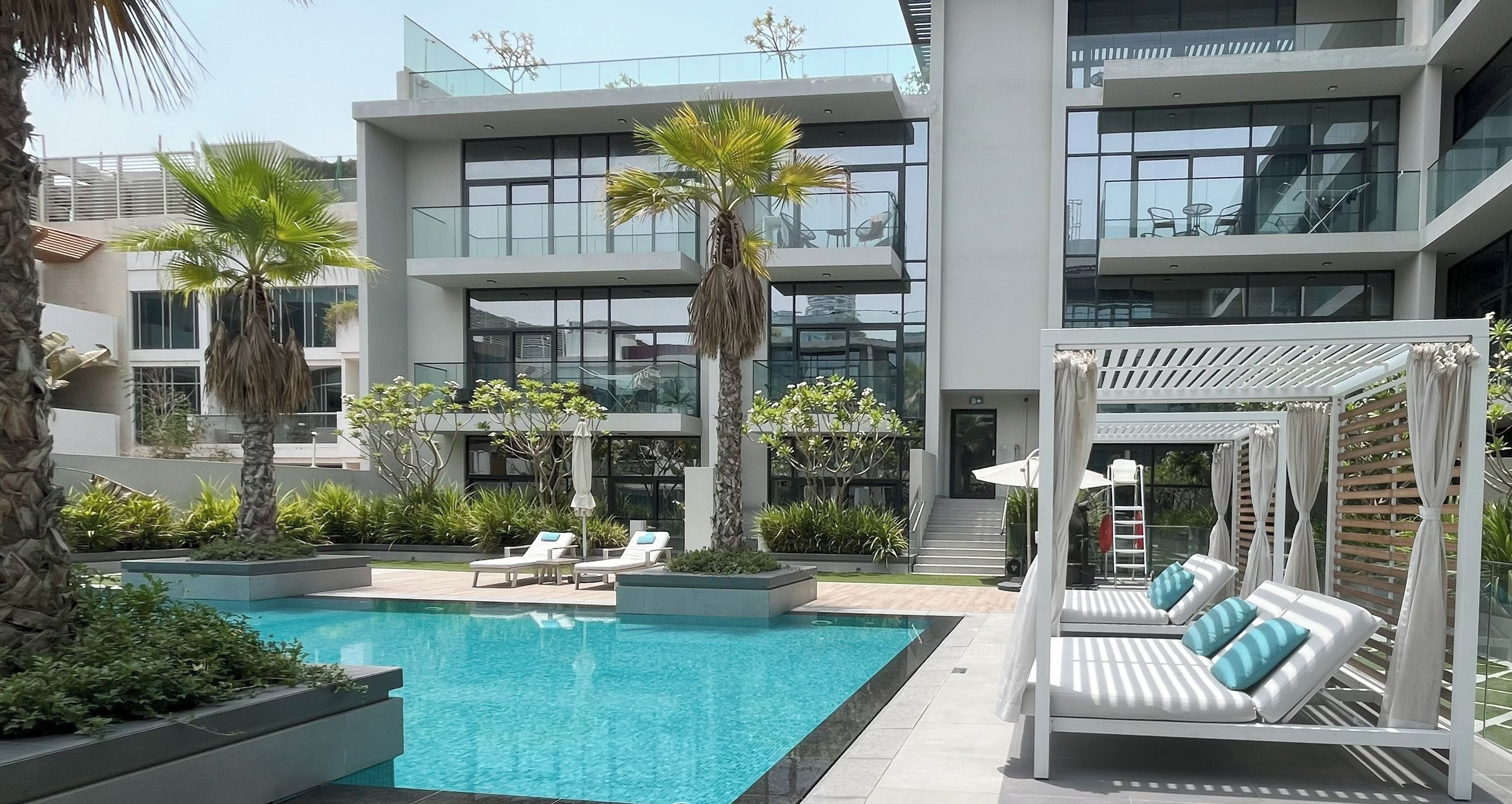 A modern building with a swimming pool, lounge chairs, and a pergola with curtains.