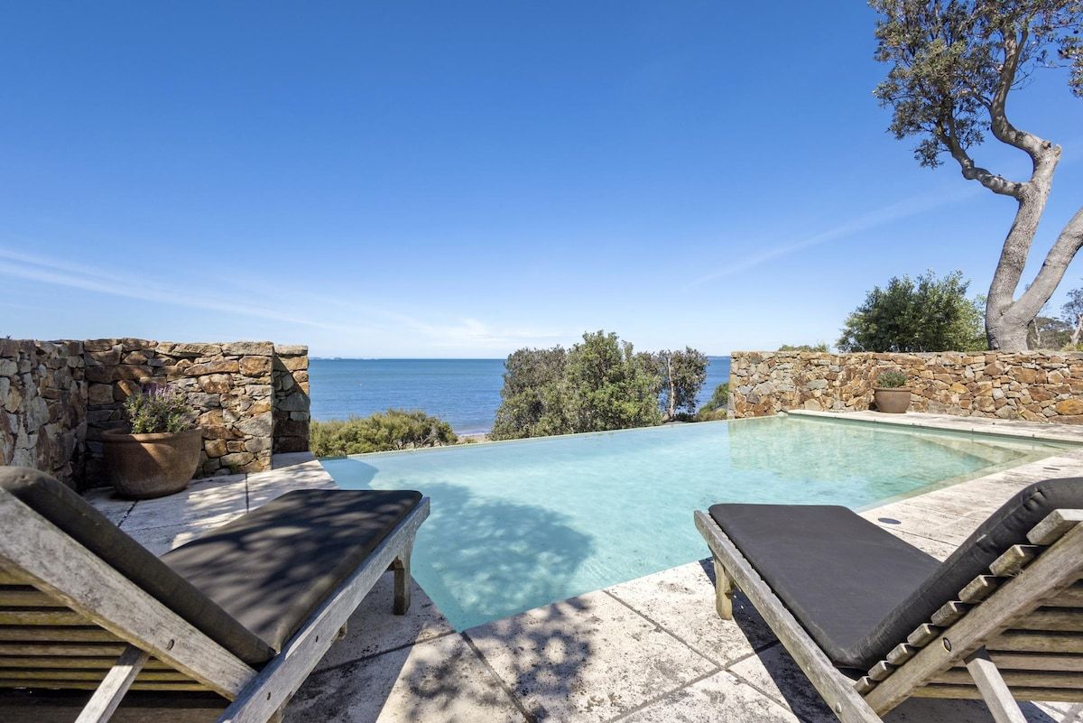 Une piscine avec deux chaises longues en bois, un mur en pierre et une vue sur la mer.