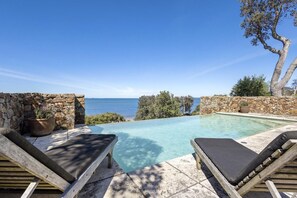 A pool with two wooden lounge chairs, a stone wall, and a view of the sea. - illiac in Somers - absolute beachfront. (Somers)