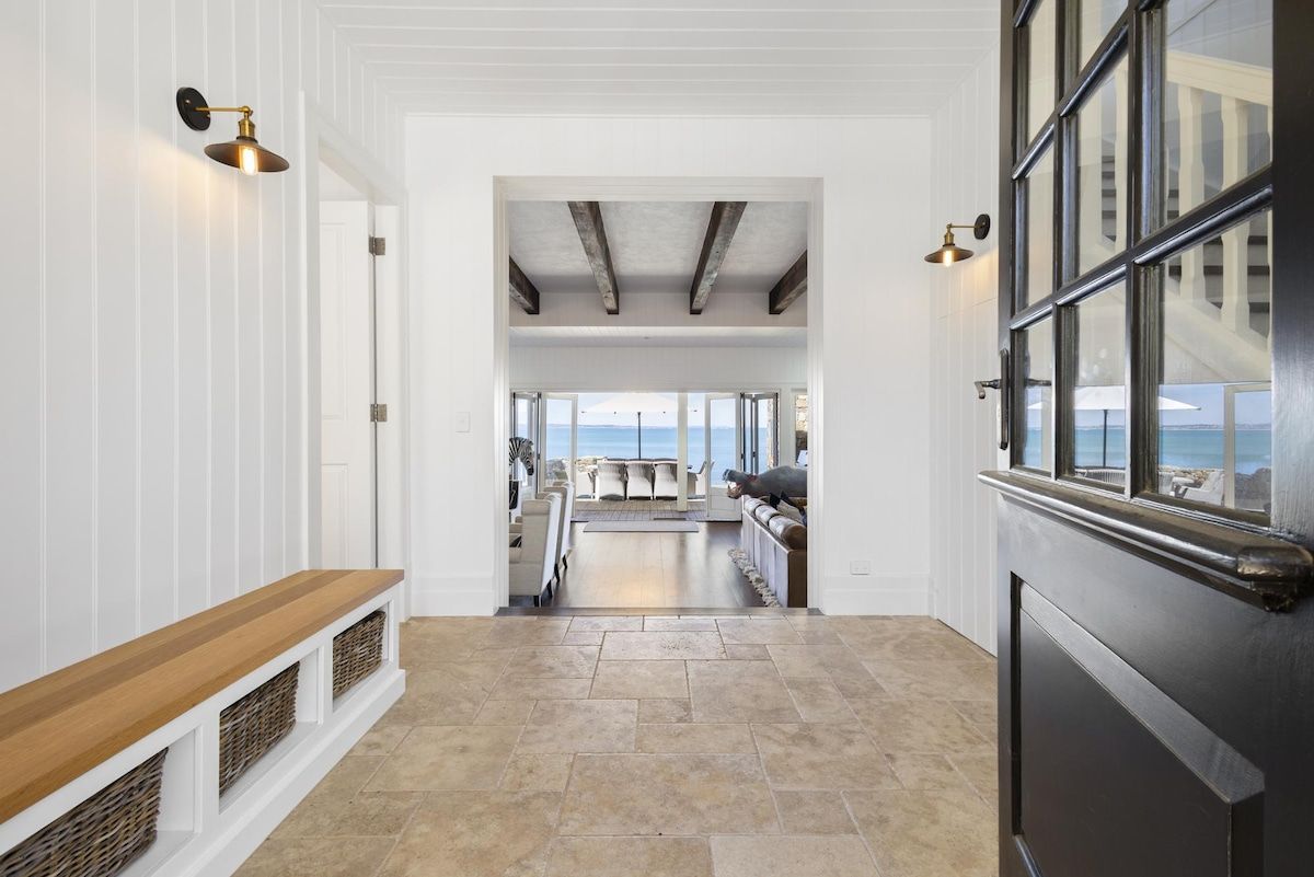 Un corridoio che conduce a una zona pranzo con vista sul mare, con una panca di legno, pavimento in pietra e una finestra con cornice nera.