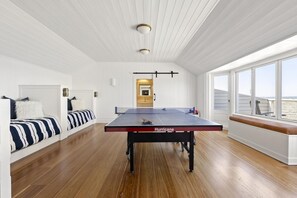 Une pièce spacieuse équipée d’une table de ping-pong, dotée d’un parquet, de murs blancs et de grandes fenêtres offrant une vue dégagée.